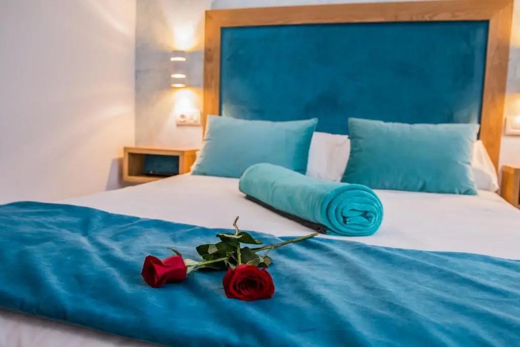 Habitación con cabecero de terciopelo azul, cojines turquesa y dos rosas rojas sobre la colcha